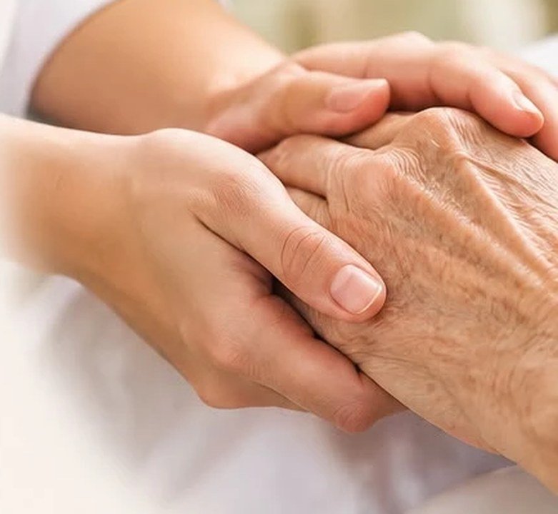 Mãos de uma cuidadora acolhendo a mão de uma pessoa idosa
